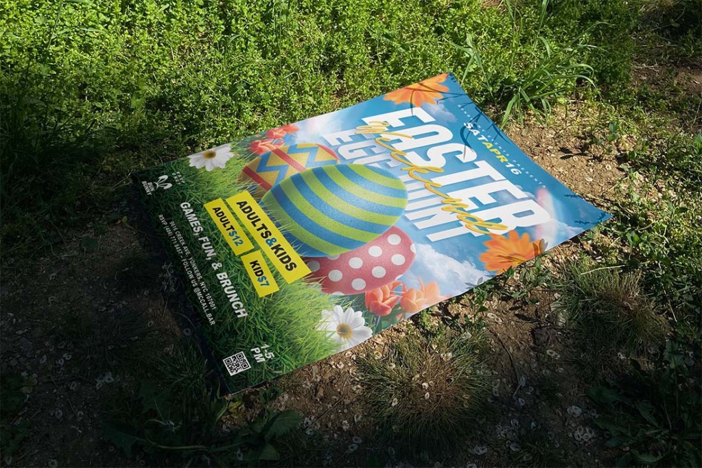 Easter Egg Hunt PSD flyer template displayed as a printed flyer mockup lying on real grass outdoors, showing the colorful 3D egg design, spring flower elements, and event info layout — CMYK print-ready Photoshop template by Bornx Design